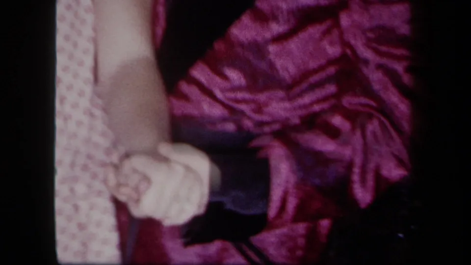 1958 New York 8mm frame with clasped hands near purple fabric and a patterned blouse in an indoor family scene.
