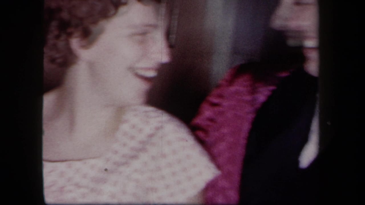 1958 New York home-movie frame with a smiling woman in a light patterned dress beside a partner partly hidden by dark clothing and magenta fabric.