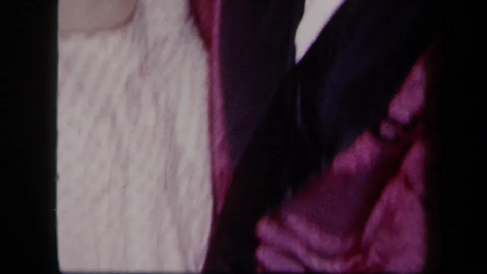 1958 New York 8mm frame showing two adults close to the camera, with clasped hands, a patterned dress, and dark and magenta clothing.