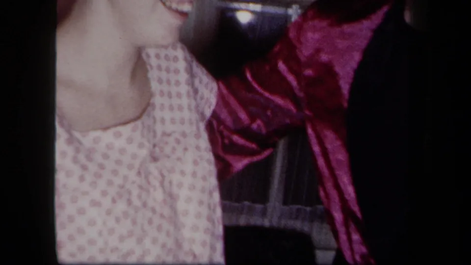 1958 New York home-movie frame showing a woman in a patterned dress close to a dance partner, with a shiny magenta garment prominent at right.