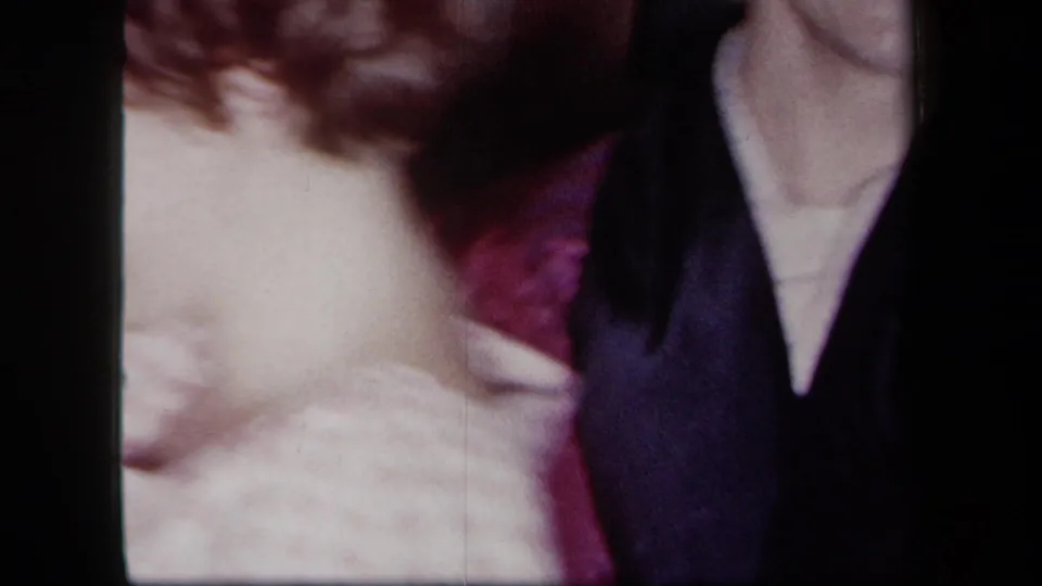 1958 New York home-movie frame showing a woman in a light dress dancing with a man in dark clothing and maroon fabric visible nearby.