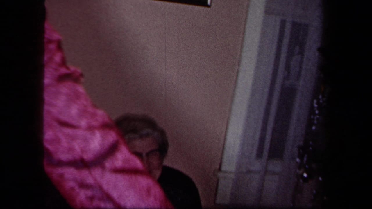 1958 New York home-movie frame with a pink-clad guest partly blocking a seated older man near a doorway in a private room.