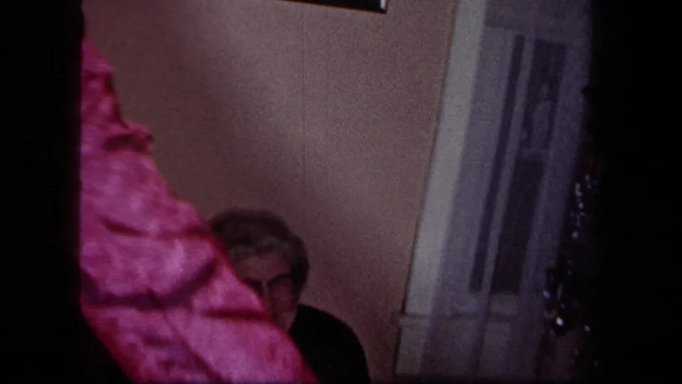 1958 New York home-movie frame with a pink-clad guest partly blocking a seated older man near a doorway in a private room.