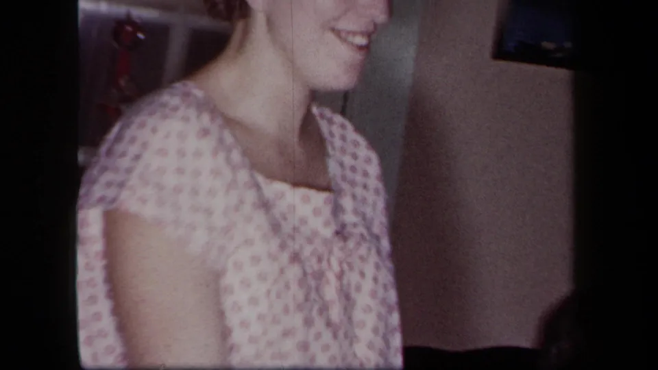 Smiling woman in a light pink patterned dress appears in a 1958 New York private interior, cropped by the handheld home-movie frame.
