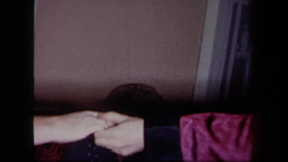 Hands meet near the lower edge of a 1958 New York home-party frame, with a dark jacket, pink sleeve, wall, and doorway visible.