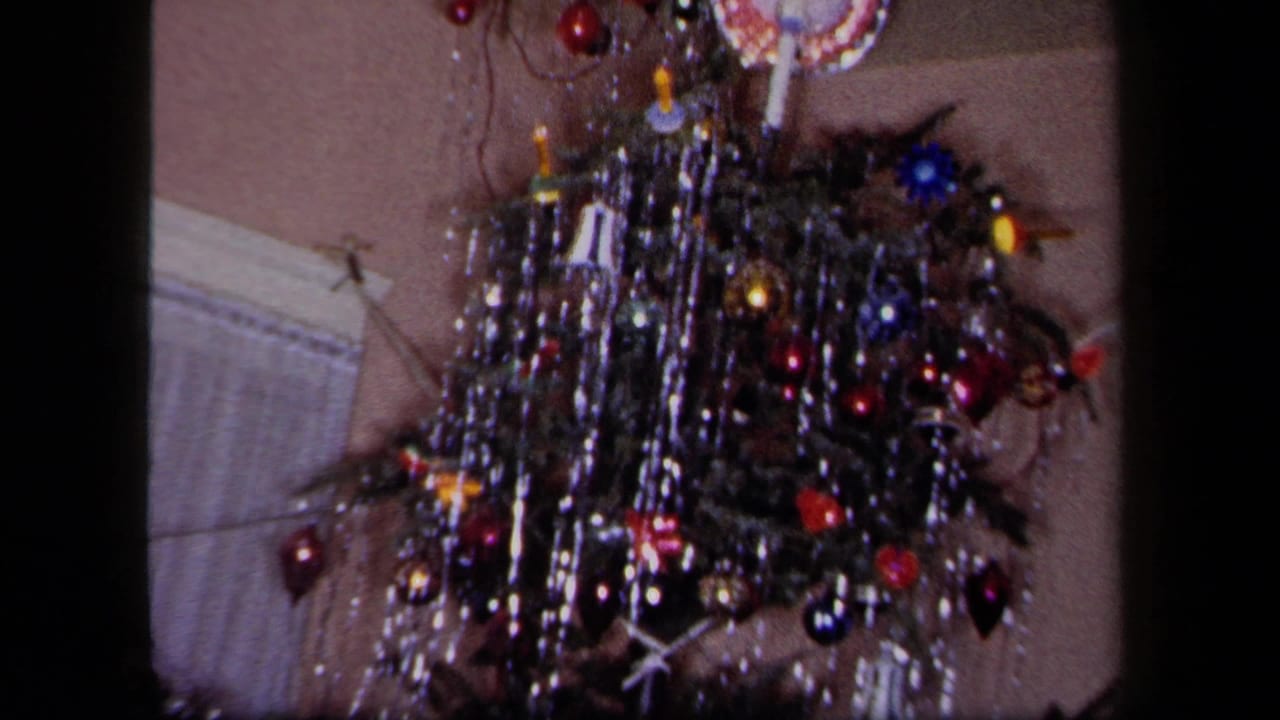 Close view of a tinsel-covered Christmas tree with colored ornaments beside a curtain.