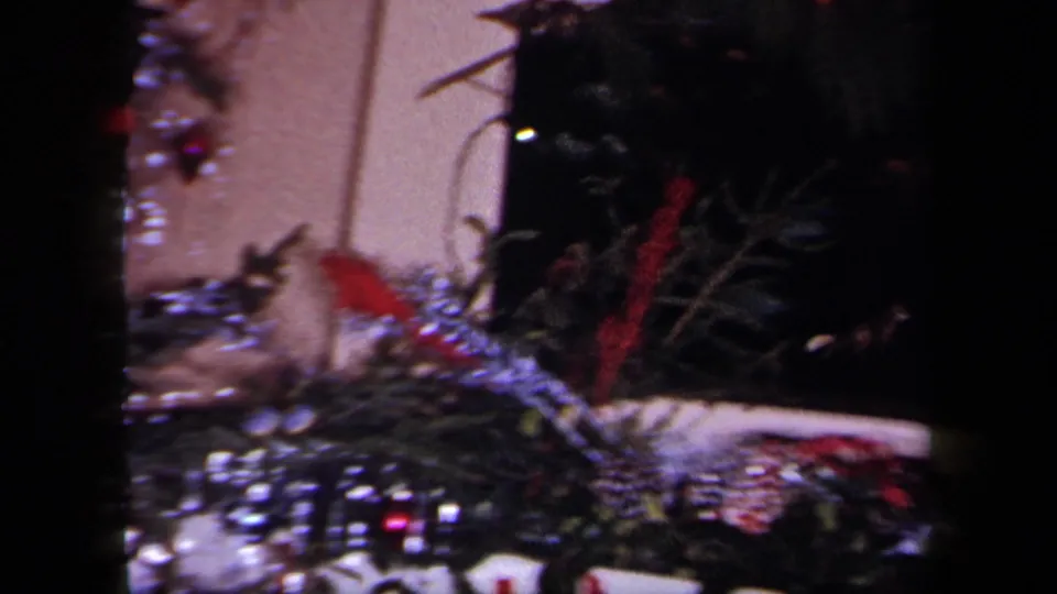 Dark 1958 New York home-movie frame showing Christmas tree branches, silver tinsel and red decorations, with an indistinct visible figure or object nearby.