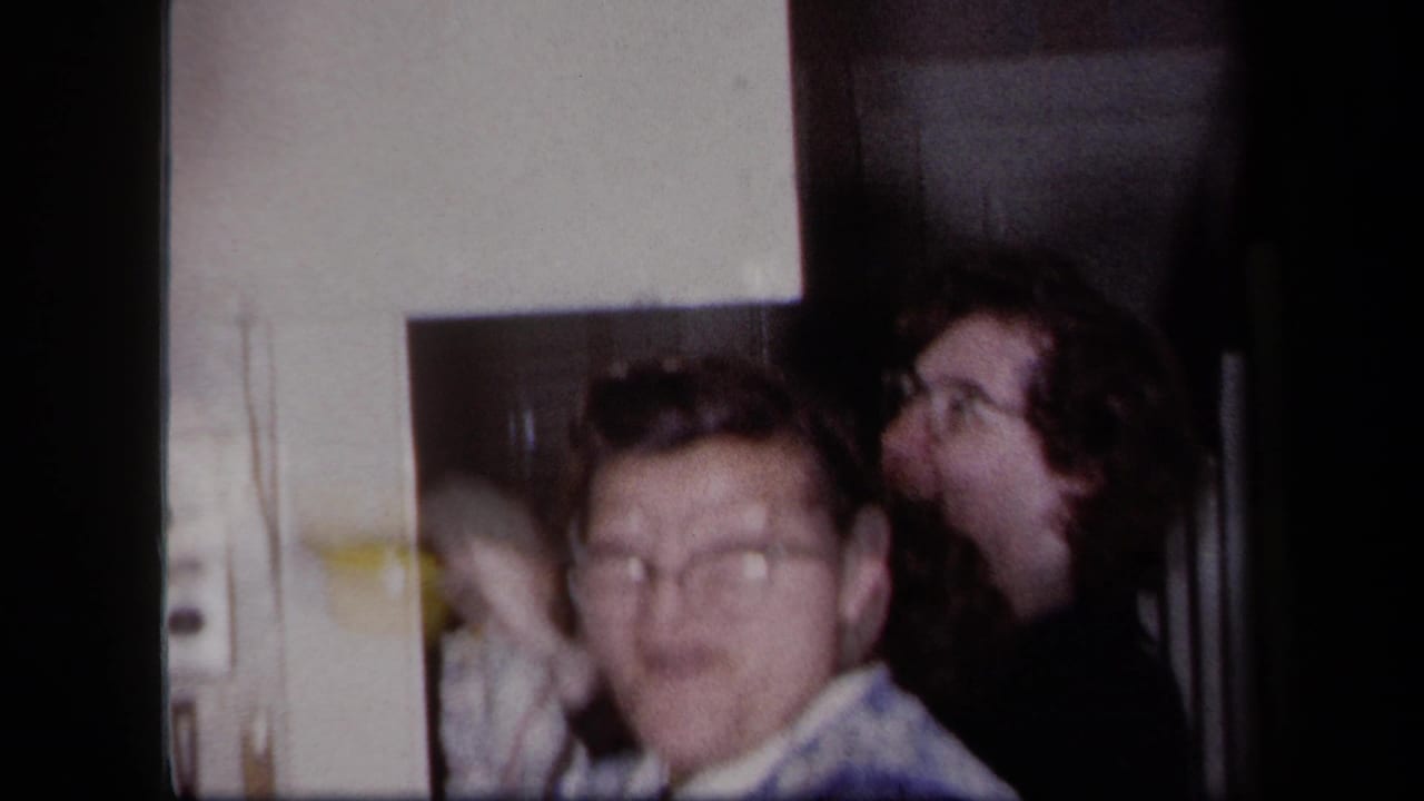 Two adults in a dim interior, one facing the camera and another turned sideways.