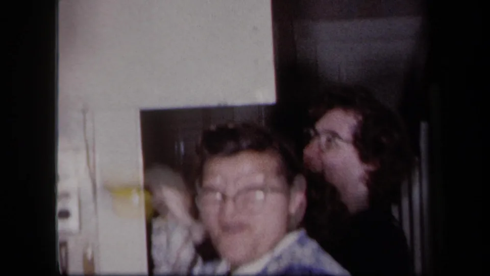 Two adults appear in a private New York interior, with one face close to the camera and another person standing behind.