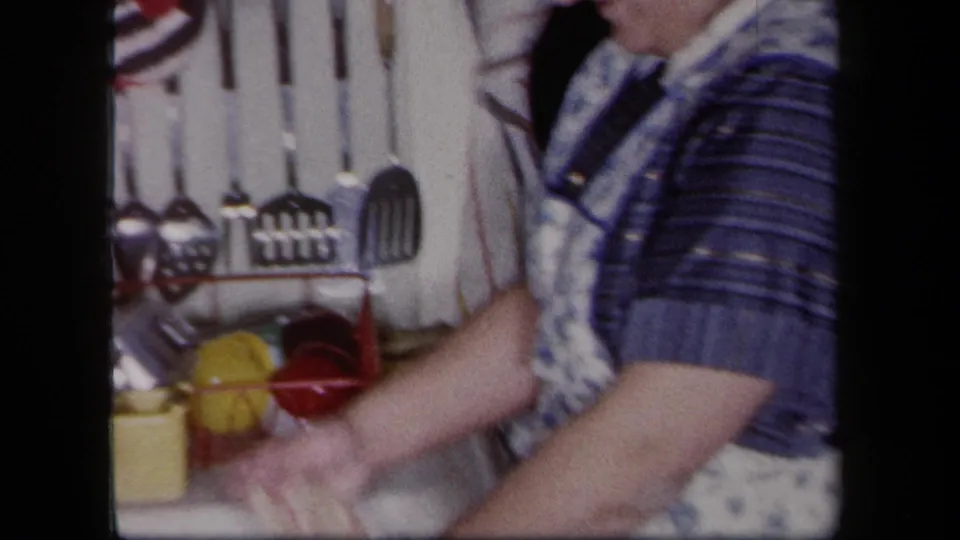 Woman in blue patterned dress and apron-like garment washes dishes in a 1958 New York kitchen with utensils on the wall.