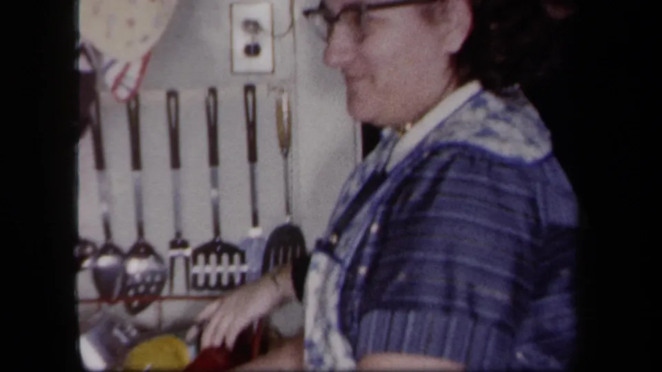A woman in glasses washes dishes in a vintage New York kitchen, with a red rack, yellow cup, and wall-mounted utensils visible.