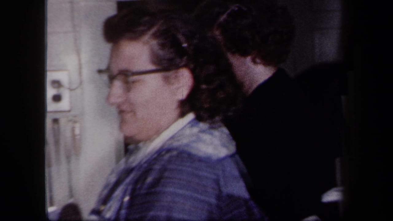 In a 1958 New York home kitchen, a woman in glasses and a blue patterned dress smiles beside a doorway.