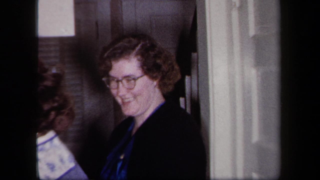 Woman with glasses smiles from a doorway as other figures stand nearby.