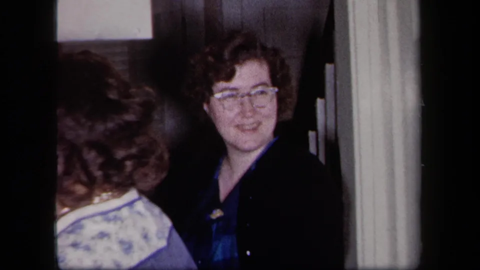 A woman with glasses looks toward another adult in a 1958 New York kitchen doorway, with soft 8mm blur.