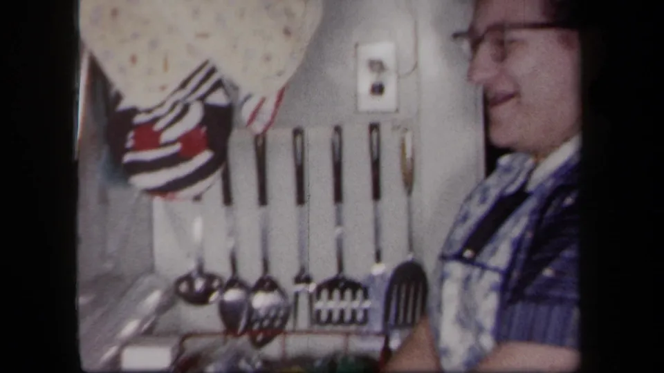 A 1958 New York kitchen wall shows hanging utensils and tiles, with a smiling woman partly visible at frame edge.
