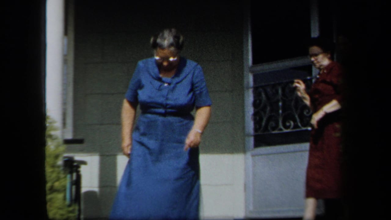 In 1958 New York home-movie footage, a woman in a blue dress steps forward from a porch as another woman in red remains near the doorway.