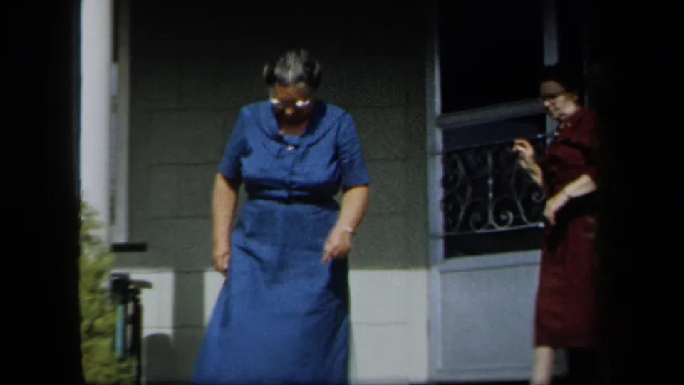 In 1958 New York home-movie footage, a woman in a blue dress steps forward from a porch as another woman in red remains near the doorway.