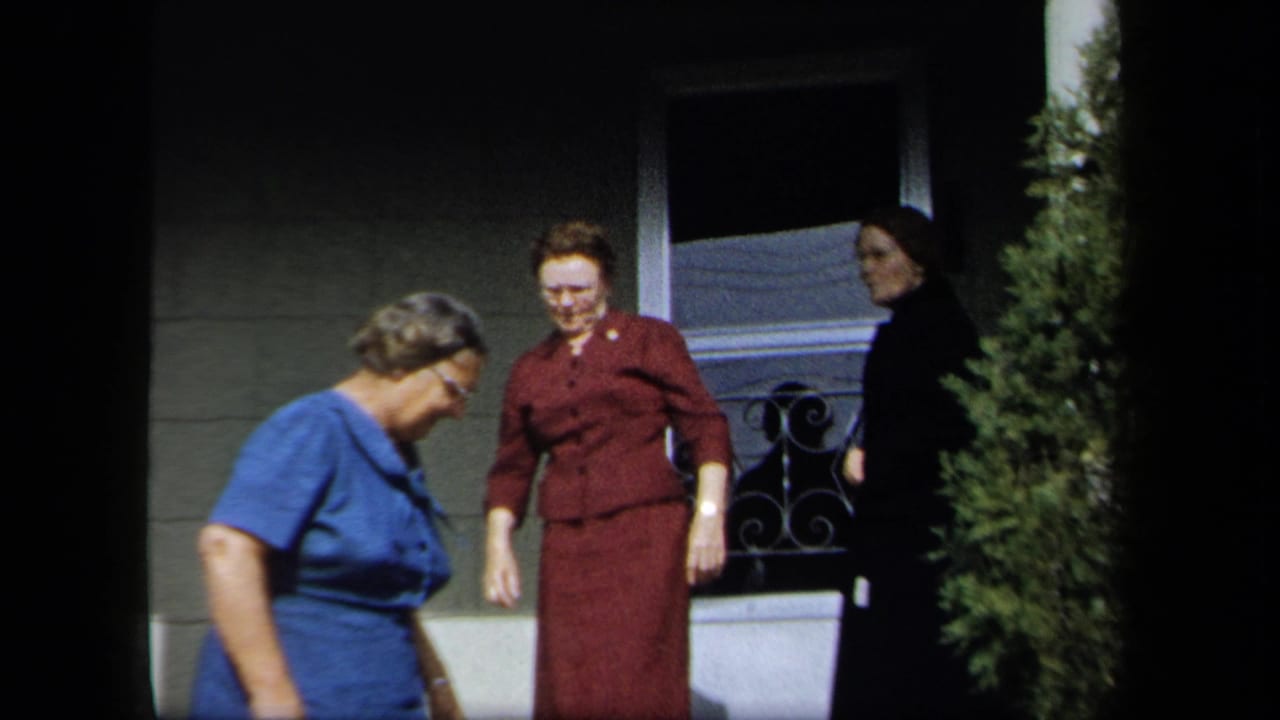 Three adult women in 1950s attire move near a residential porch in New York, with a screen door, railing, and evergreen shrub visible.