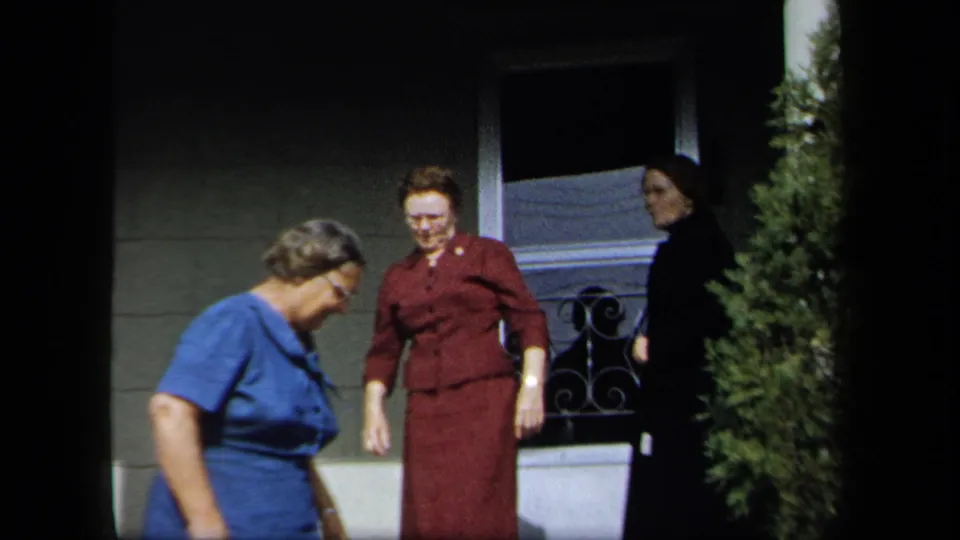 Three adult women in 1950s attire move near a residential porch in New York, with a screen door, railing, and evergreen shrub visible.