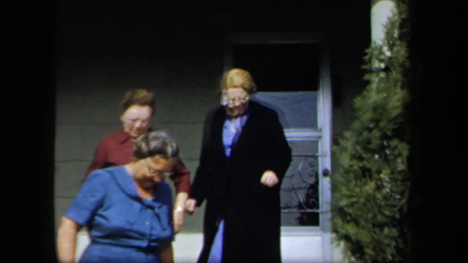 Adult women in blue, red, and dark clothing walk down from a porch area in 1958 New York home-movie footage.
