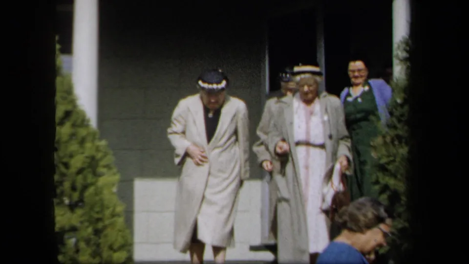 1958 New York frame showing older women in hats and light coats exiting a building, with faces visible.