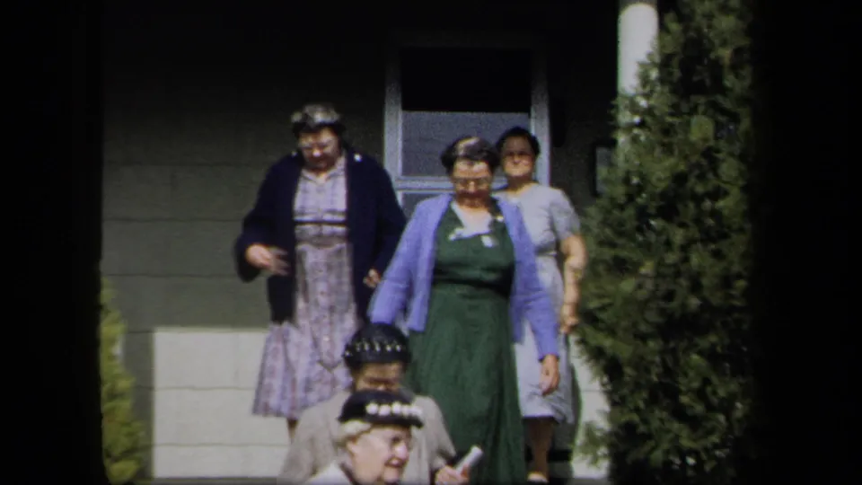 Adult women in 1950s attire move outside a New York residence, with a doorway, steps, and tall shrubbery visible.