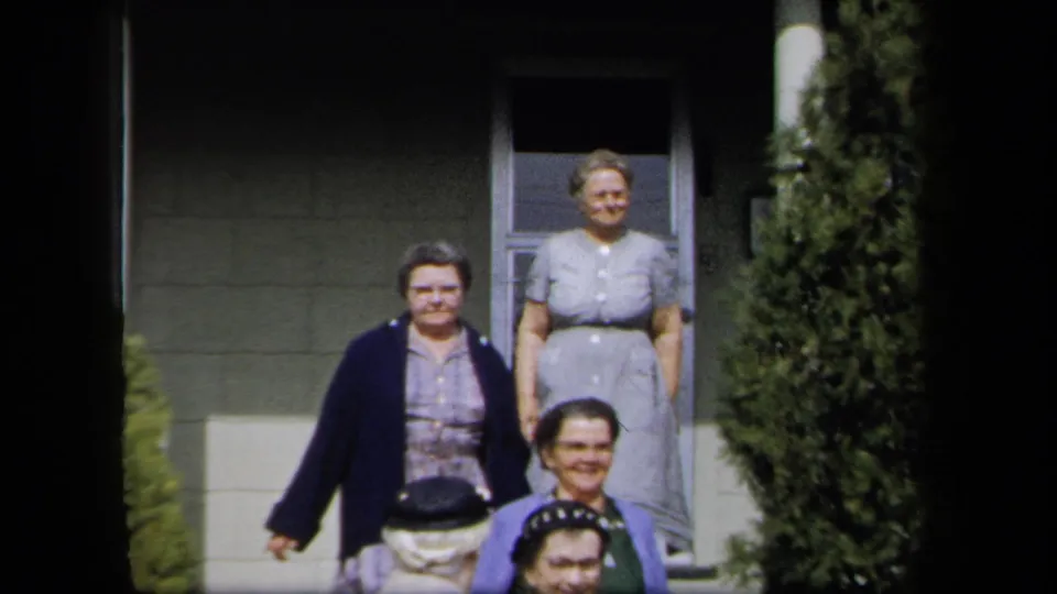 Adult women in dresses and cardigans appear outside a New York residence, framed by the doorway and shrubs in 8mm color.