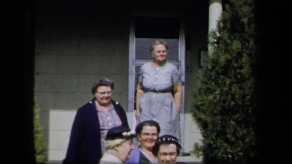 Adult women in 1950s dress gather outside a New York residence, some standing on porch steps and others partly framed below.
