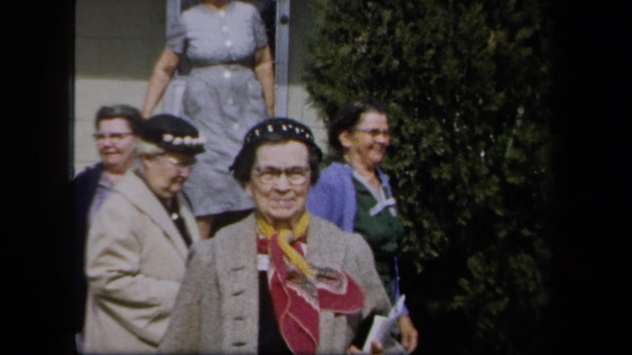Several women stand near tall shrubs, with one woman facing the camera in a hat and scarf.