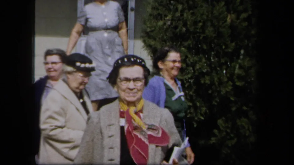Women in 1950s clothing stand outside a New York house, including a woman in a decorated hat facing the camera.