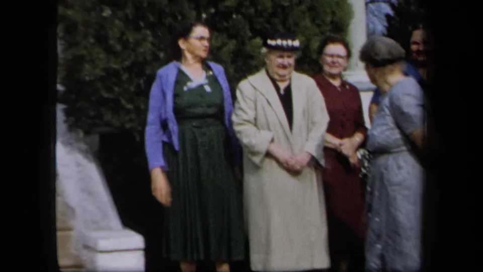 A loose group of adult women in 1950s dresses and coats gathers outside a New York residence before posing.