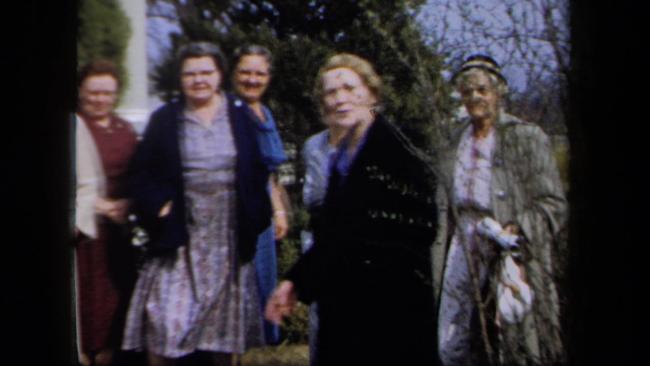 In 1958 New York home-movie footage, several women in dresses, coats, and hats gather outside near shrubs and a building entrance.