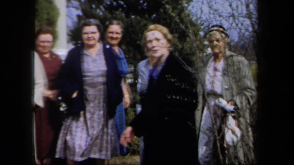 In 1958 New York home-movie footage, several women in dresses, coats, and hats gather outside near shrubs and a building entrance.