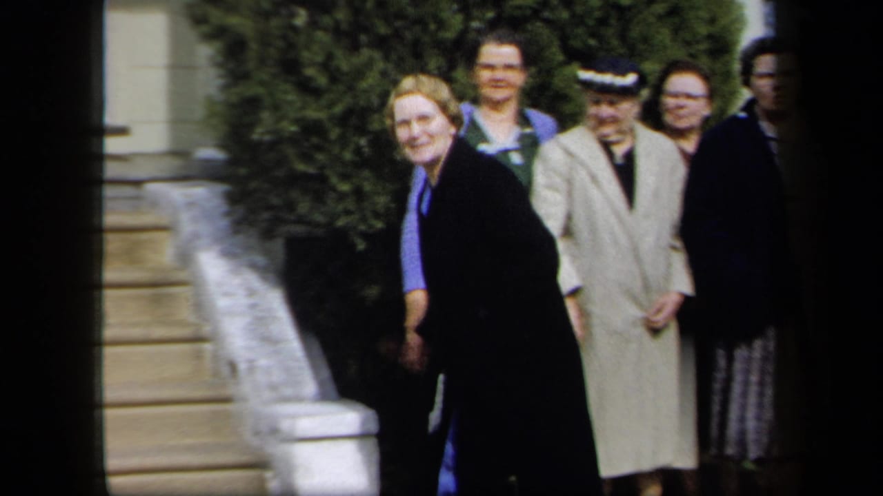 Women line up near stone steps and shrubs outside a building.