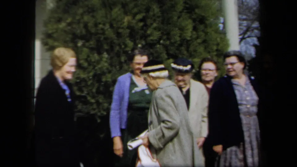 Women in coats, dresses, and hats shift positions and talk near stone steps in 1958 New York home-movie footage.