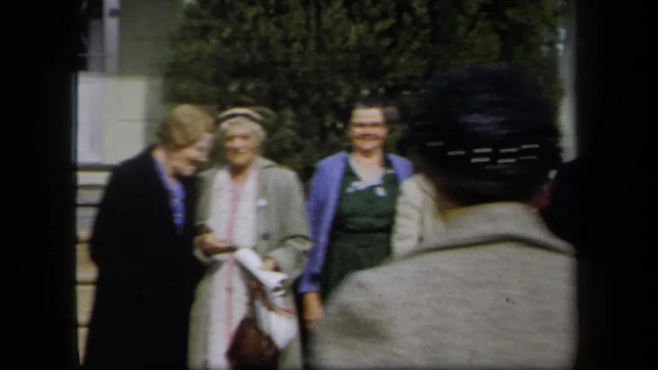 A woman in a light coat and others stand near stone steps as a foreground figure faces them in 1958 New York home-movie footage.