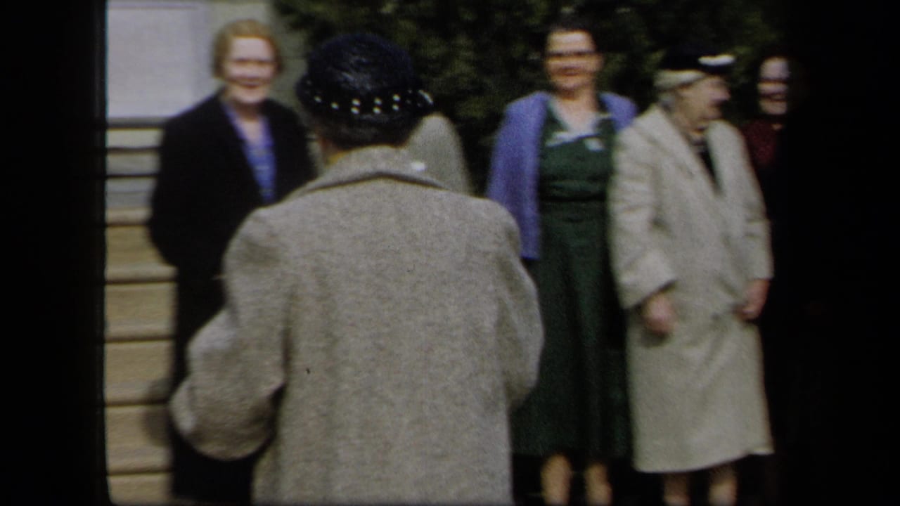 1958 New York 8mm frame showing women in mid-century clothing near porch steps as a hat-wearing woman stands with her back to camera.