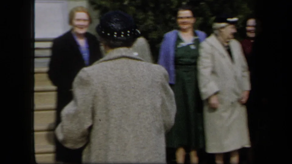 1958 New York 8mm frame showing women in mid-century clothing near porch steps as a hat-wearing woman stands with her back to camera.