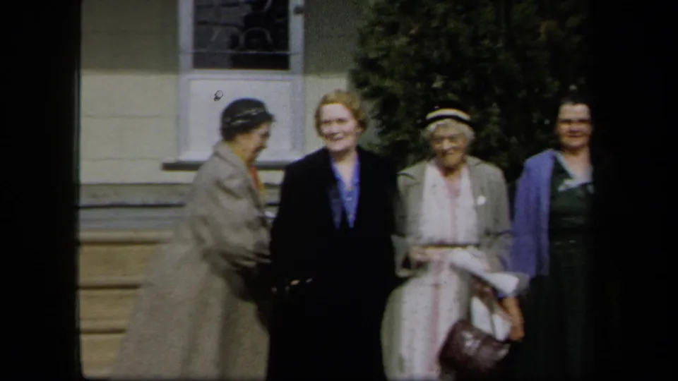 1958 New York 8mm frame showing women in coats and dresses gathered by a house exterior, smiling toward the camera.