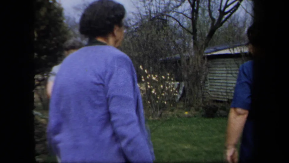 1958 New York home-movie frame showing a woman in a purple sweater walking across a yard with trees and a wooden shed behind her.