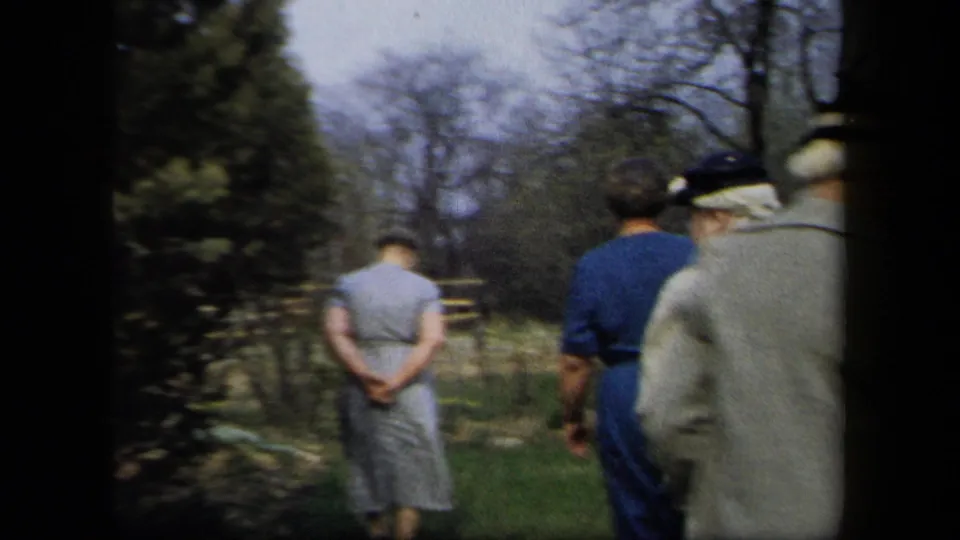 Rear-view 8mm frame of adults in coats and dresses walking through a New York garden setting in 1958.