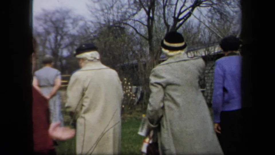 Adults in coats and hats gather near a garden fence in 1958 New York home-movie footage, seen mostly from behind.