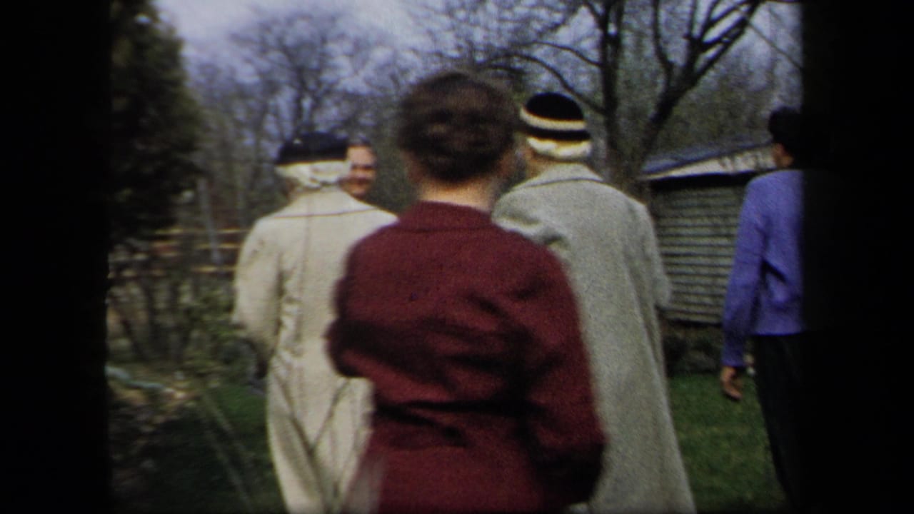 1958 New York home-movie frame showing adults in mid-century coats and hats walking away through a garden area.