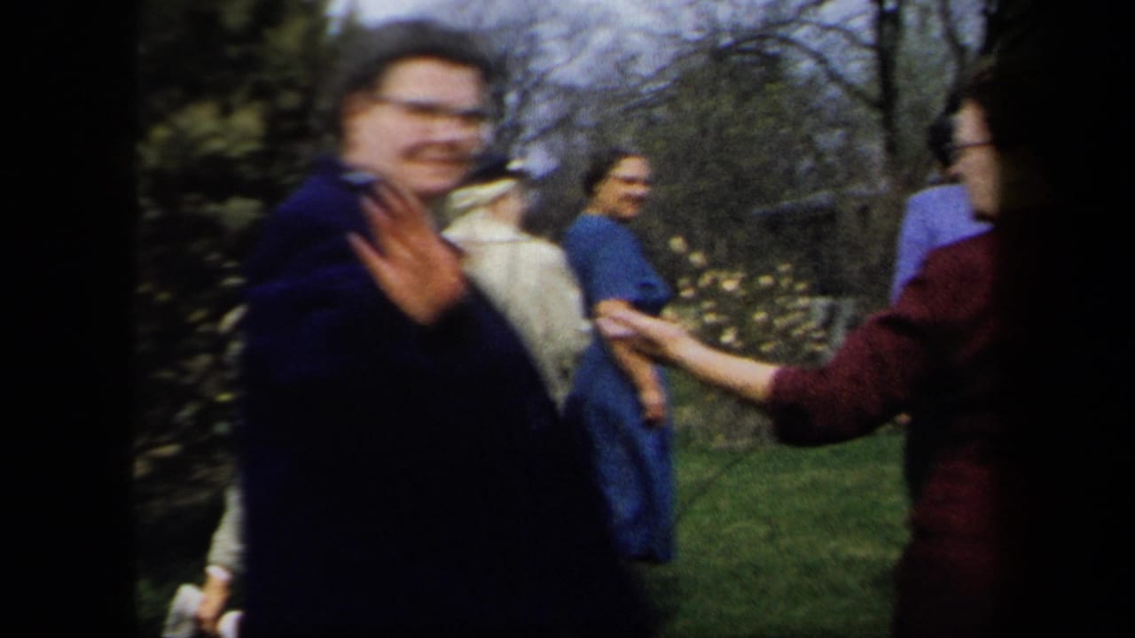 1958 New York garden scene with a man raising his hand near the camera as other adults stand or walk behind him.