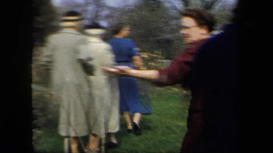 Handheld 1958 New York home-movie frame showing adults walking across grass, with one person at right extending an arm.