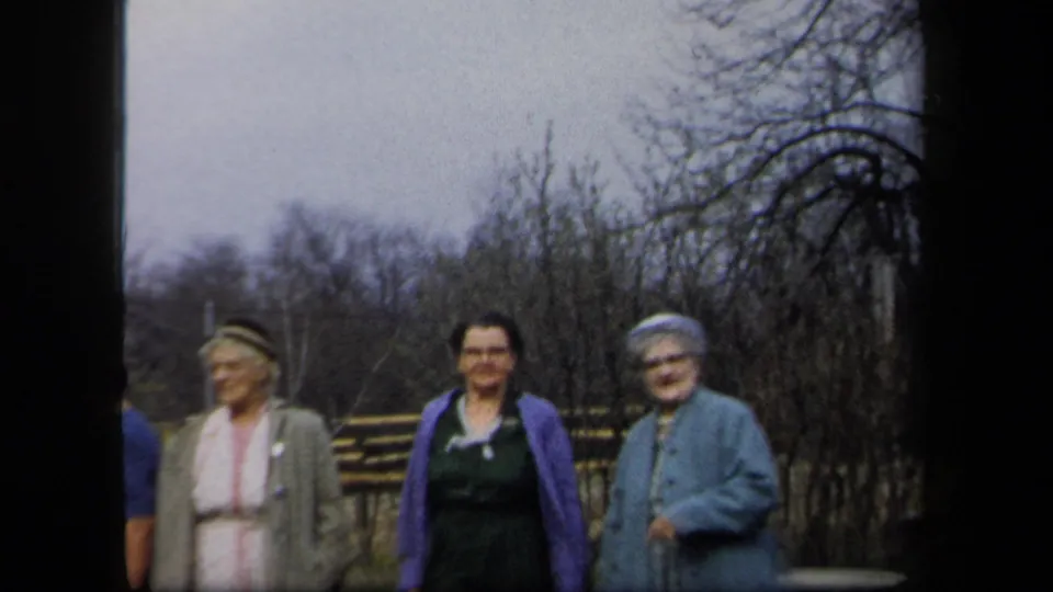 1958 New York home-movie frame showing three women in coats and dresses standing together in a garden setting.