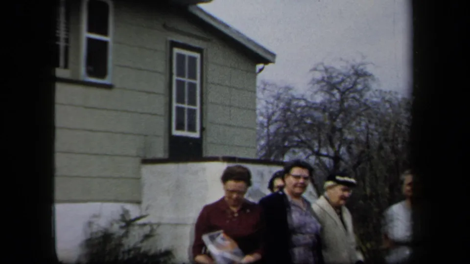 Adult women in period clothing stand beside a residential house in the New York 1958 home-movie footage.