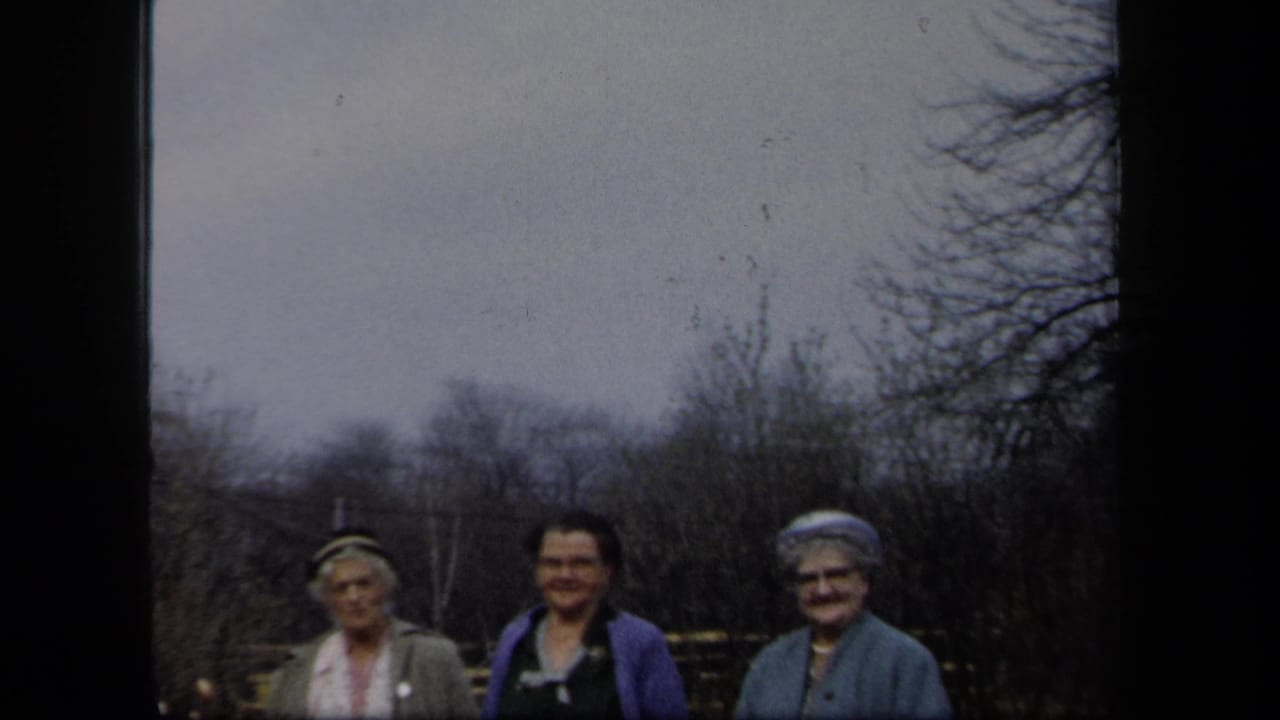 Three adult women in period clothing smile outdoors in New York, framed by bare trees, garden fencing, and 8mm film borders.