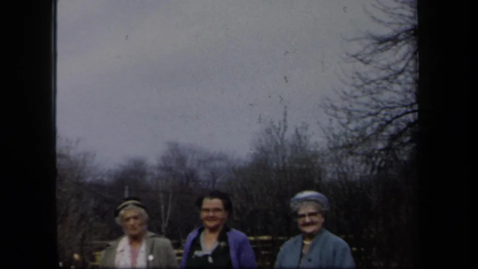 Three adult women in period clothing smile outdoors in New York, framed by bare trees, garden fencing, and 8mm film borders.