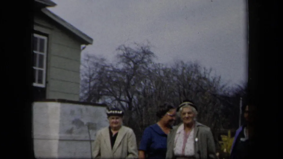 Women in 1950s attire stand near a house exterior and garden wall in New York, with bare trees and soft 8mm color visible.